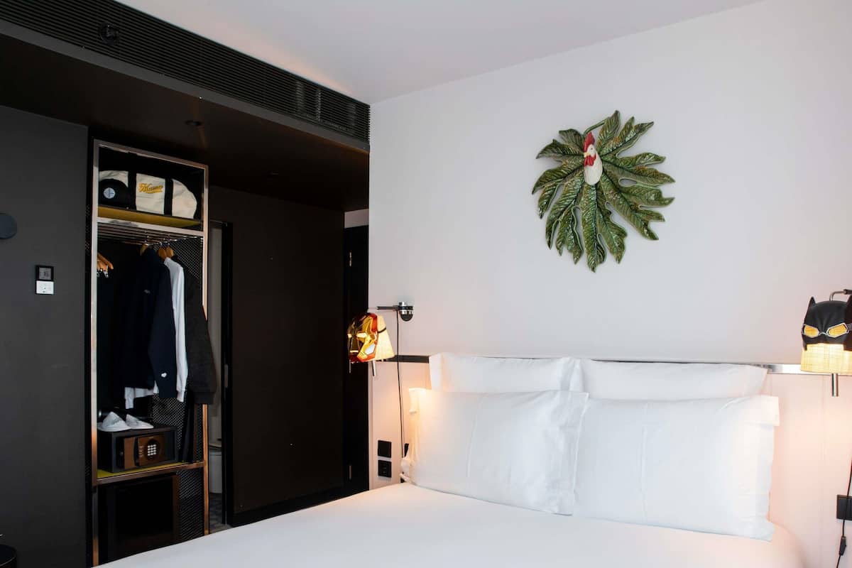 A hotel room with a bed, a wardrobe, a coat rack, a wall-mounted lamp, and a decorative leaf wall piece.