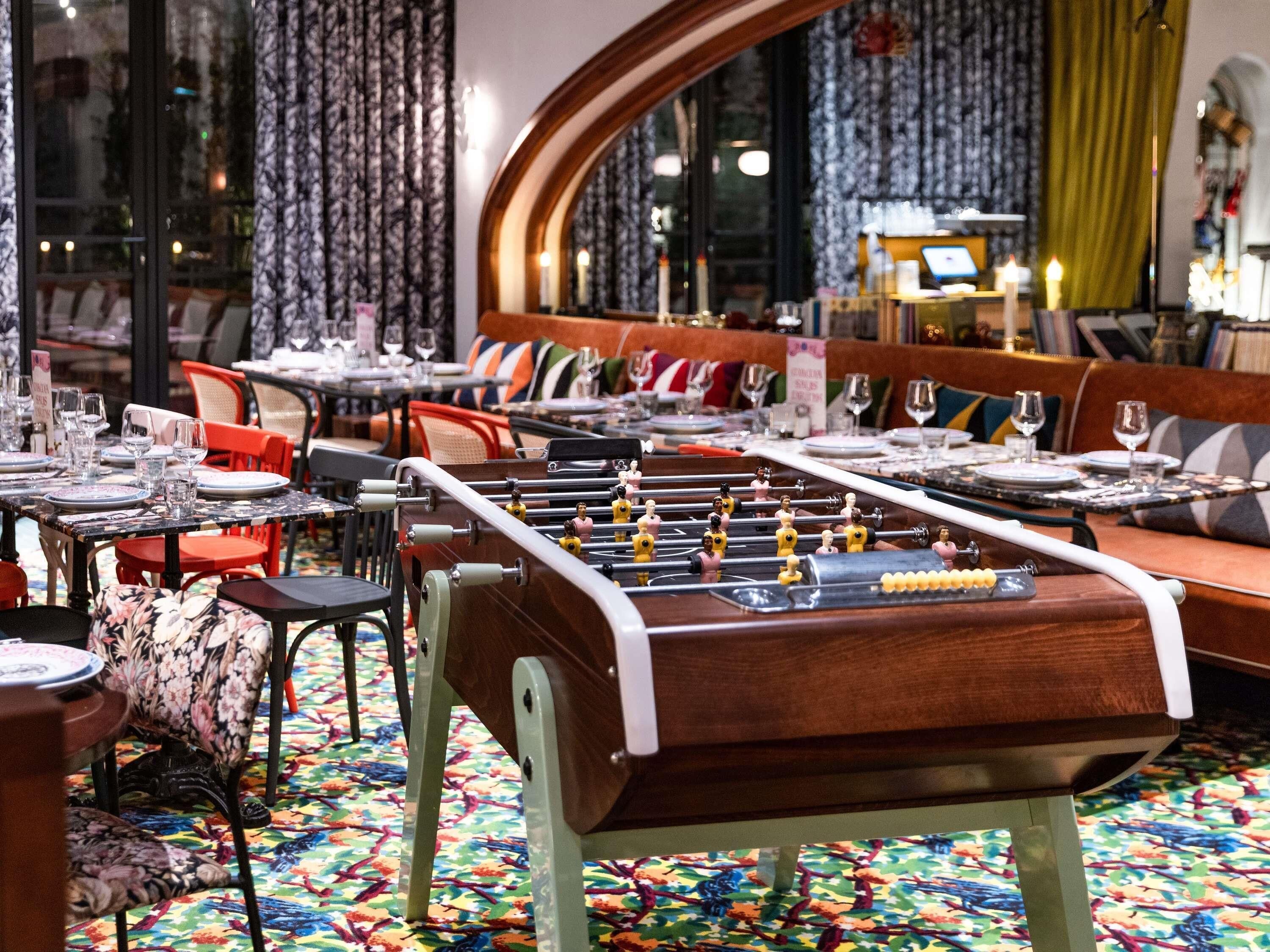 A restaurant with tables set for dining, a foosball table, and a colorful carpet.
