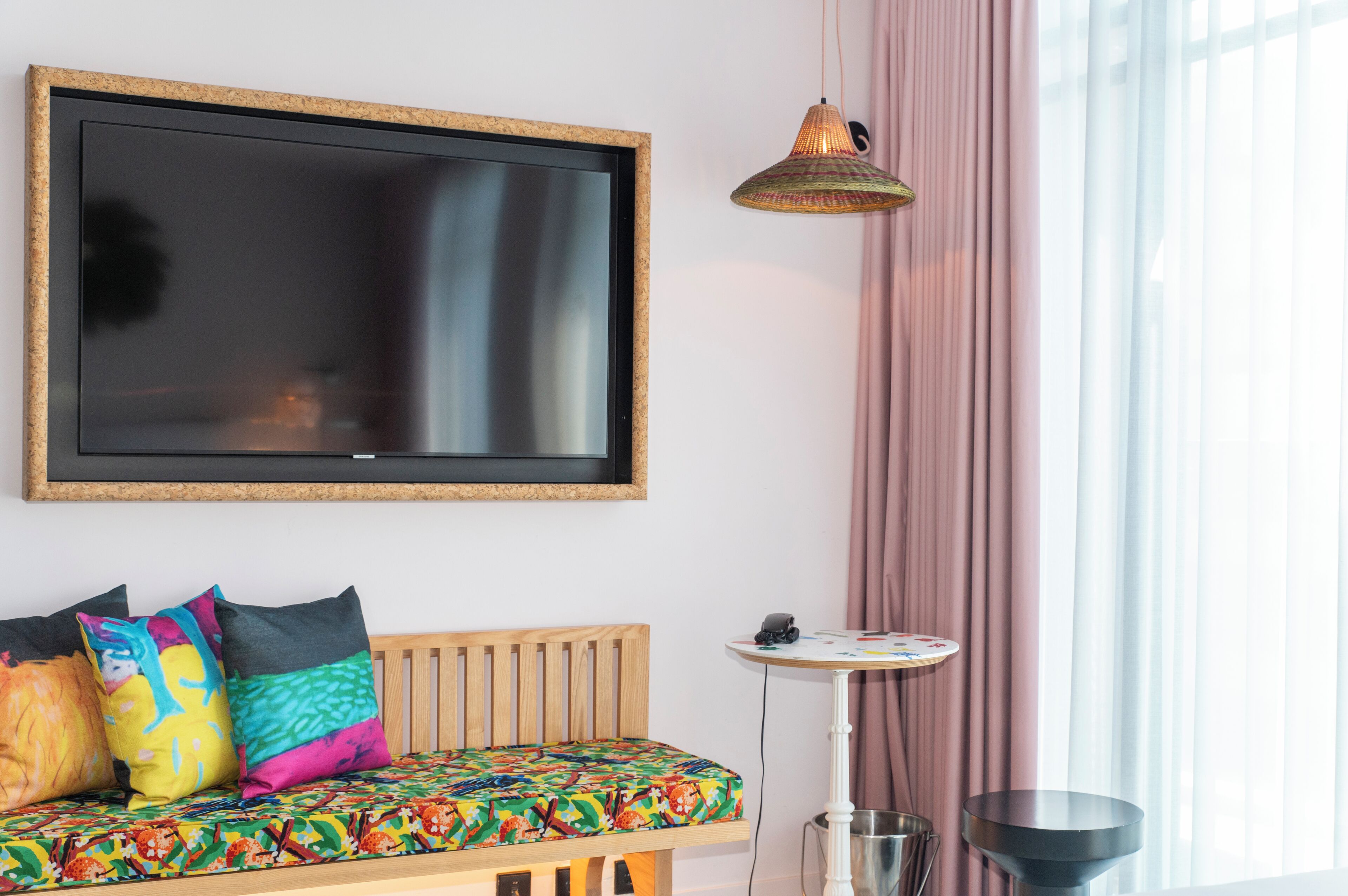 A room with a large flat-screen TV mounted on the wall, a wooden bench with colorful cushions, a small round table, and a hanging lamp.