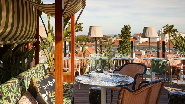 An outdoor seating area with tables and chairs, a striped canopy, and a view of buildings in the background. - Mama Shelter Lisboa  (Lisbon)