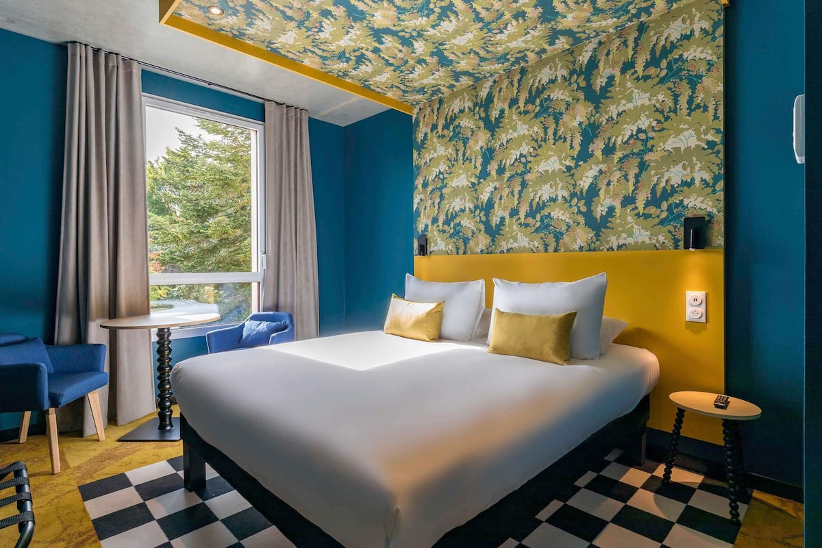 Una habitación de hotel con una cama grande, una mesita, una silla, un ventanal con cortinas y un techo con diseño.