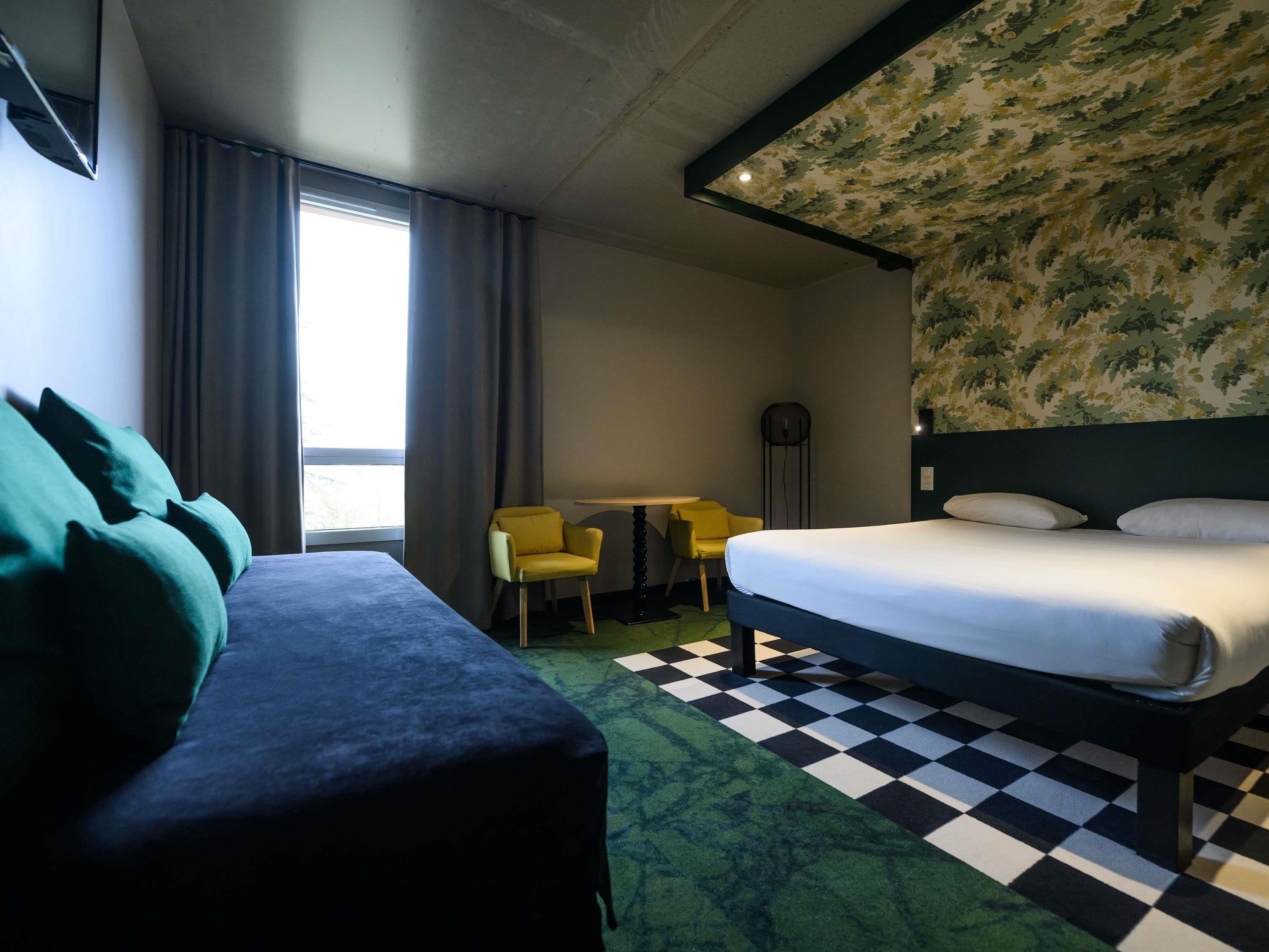 A hotel room with a bed, a sofa, a desk with chairs, a floor lamp, and a patterned wall.