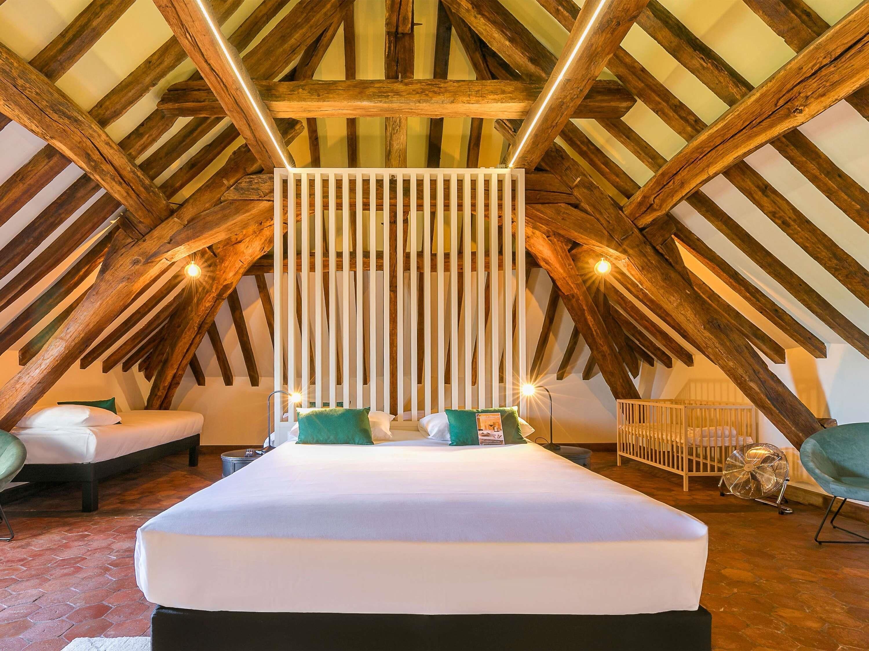 A bedroom with a sloped ceiling, wooden beams, a bed with white bedding, a white partition, and a fan.