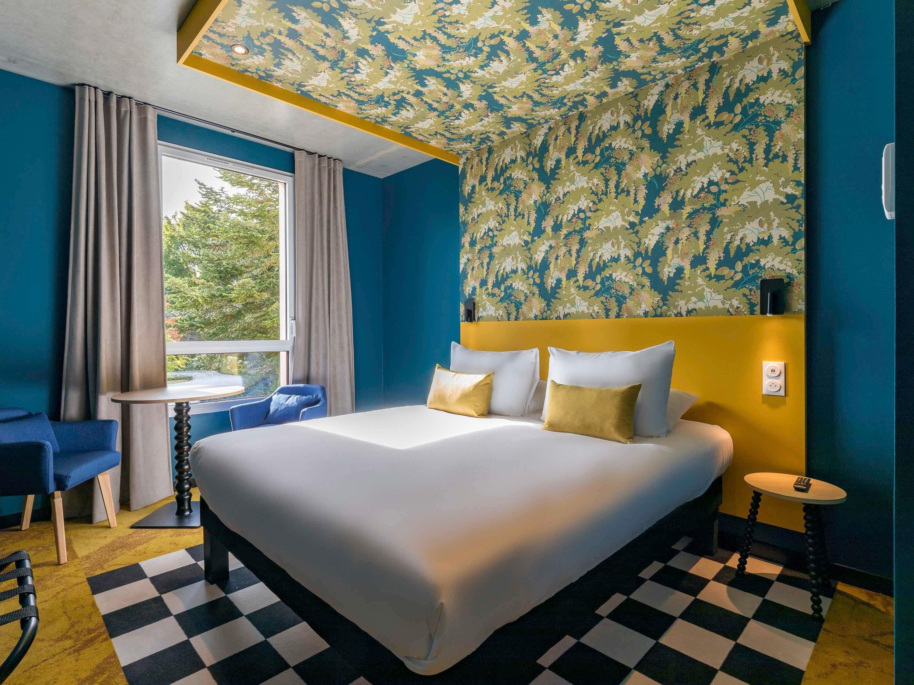 A hotel room with a large bed, a small table, a chair, a window with curtains, and a patterned ceiling.
