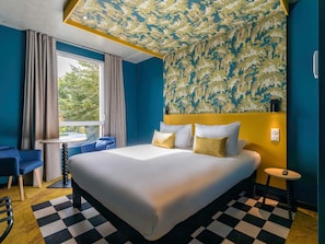 A hotel room with a large bed, a small table, a chair, a window with curtains, and a patterned ceiling. - ibis styles Dijon Sud Domaine de Beauregard (Longvic)