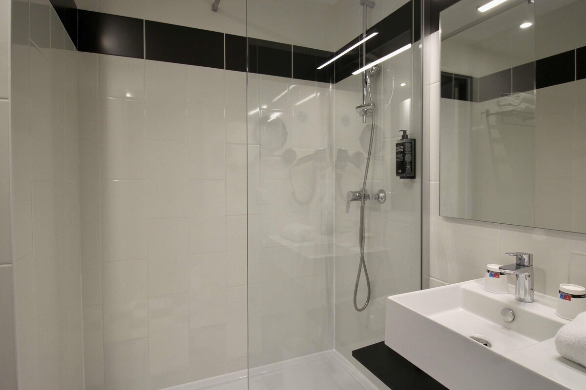 Een moderne badkamer met een douchecabine van glas, een witte wastafel met een zwart aanrechtblad en een grote spiegel boven de wastafel.