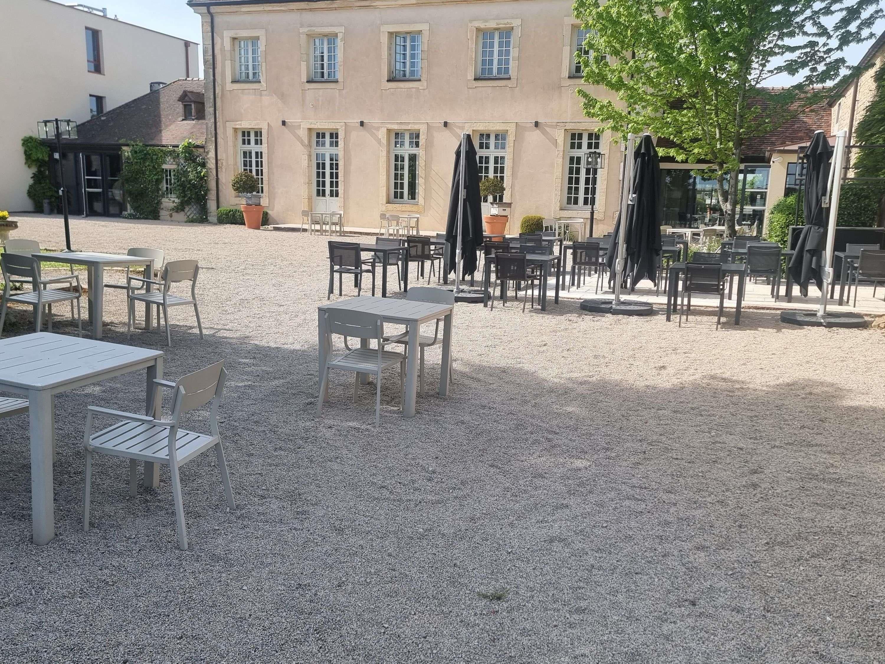 An outdoor seating area with tables and chairs, surrounded by a gravel surface, and a building with multiple windows in the background.