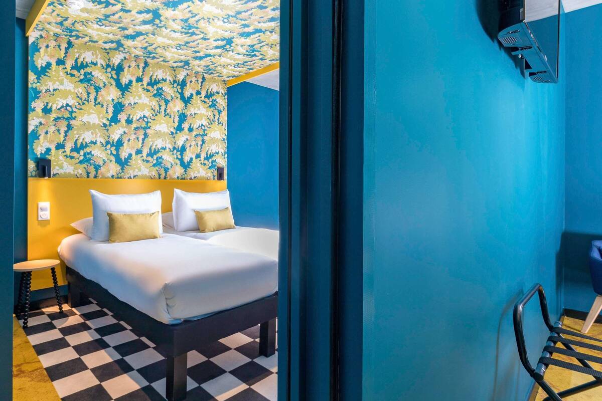 Una habitación de hotel con piso a cuadros, una cama con sábanas blancas y dos almohadas, una mesita de noche pequeña y una pared con estampado floral.