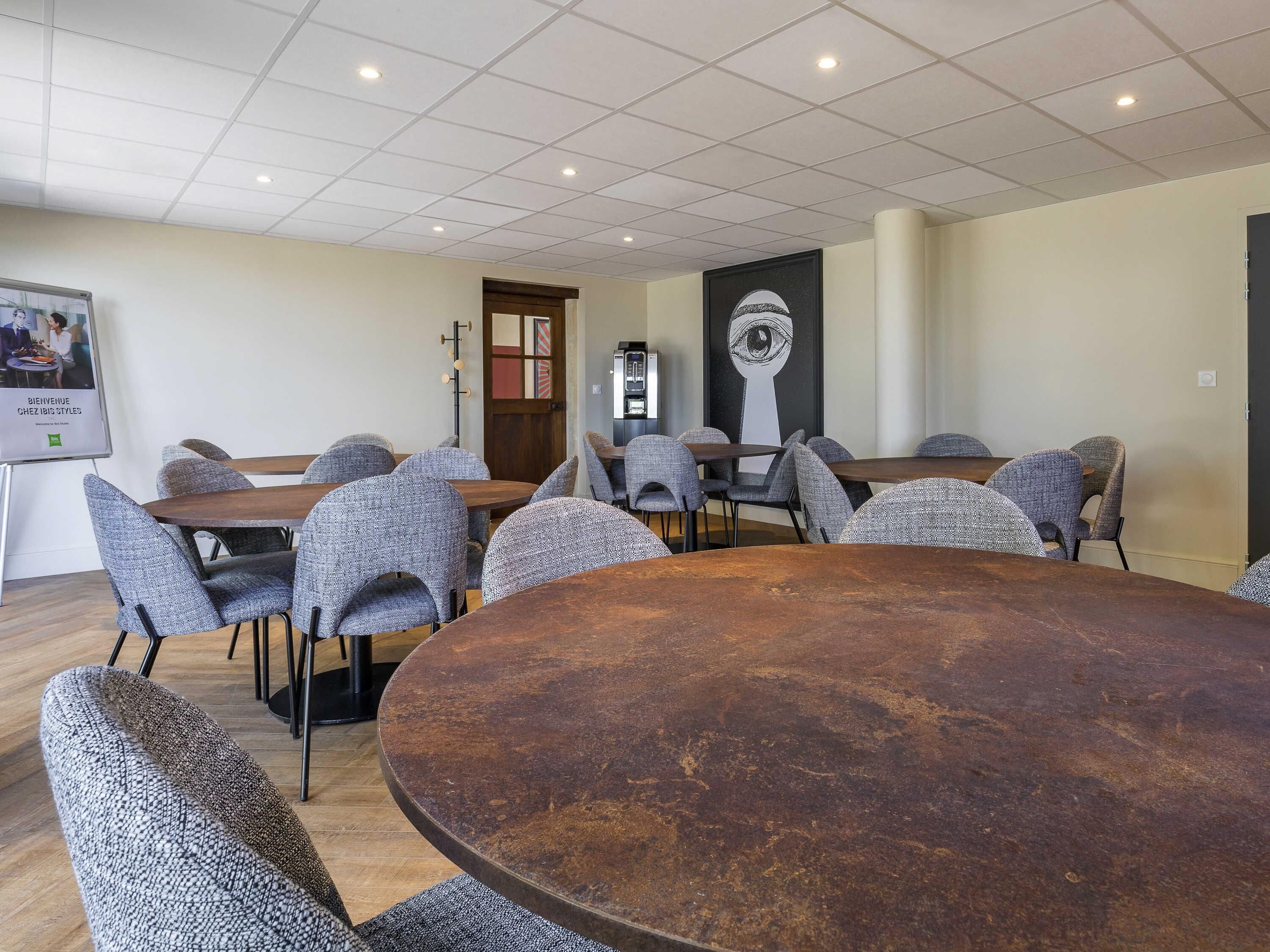 A conference room with round tables and grey upholstered chairs, a projector screen, and a wall-mounted clock.
