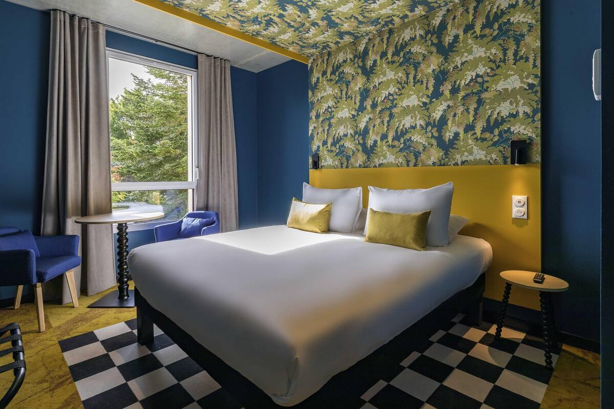 A hotel room with a bed, a desk, a chair, and a view of trees outside.