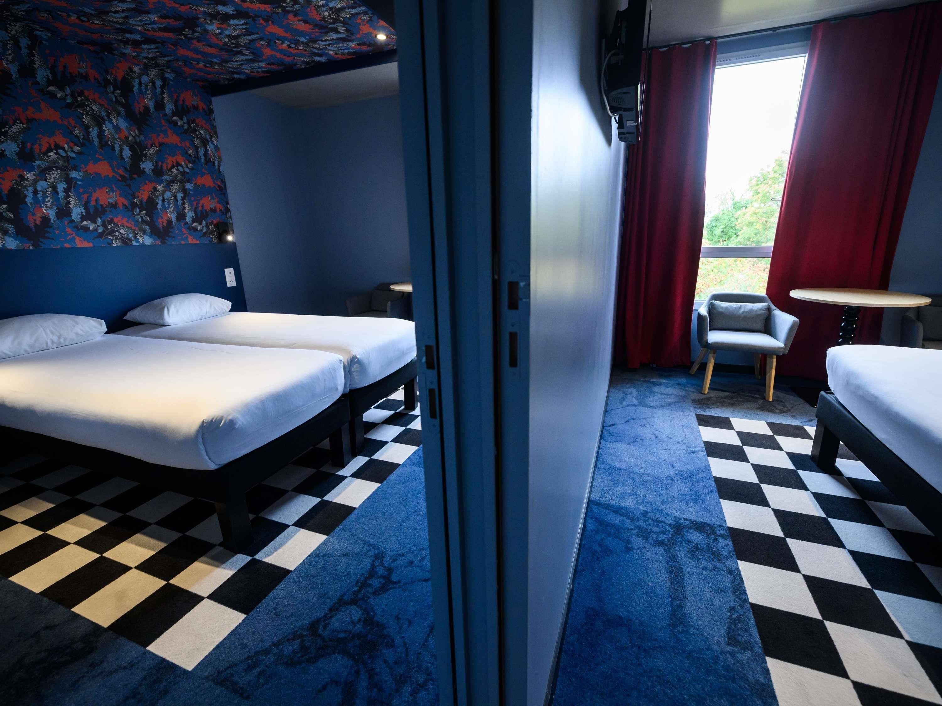 A hotel room with two beds, a small table, a chair, and a view of greenery outside.
