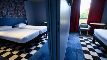 A hotel room with two beds, a small table, a chair, and a view of greenery outside.