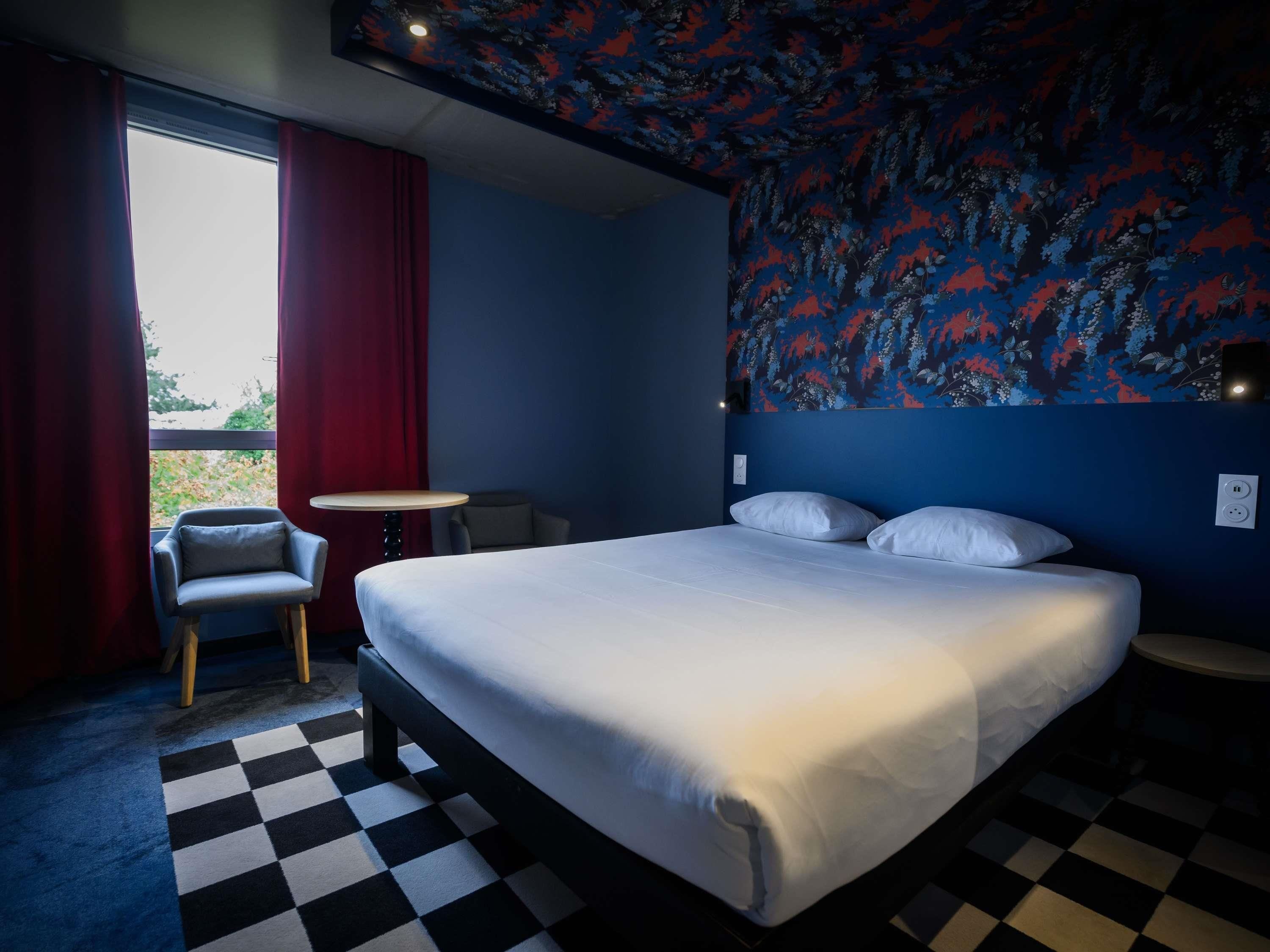 A hotel room with a large bed, a small table, a chair, and a window with curtains.
