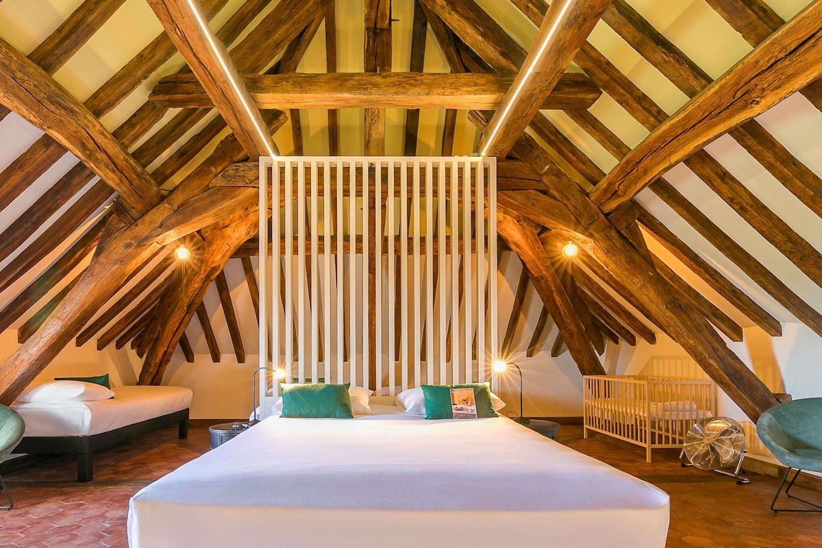 Een slaapkamer met een schuine kap, houten balken, een bed met wit beddengoed, een witte scheidingswand en een ventilator.