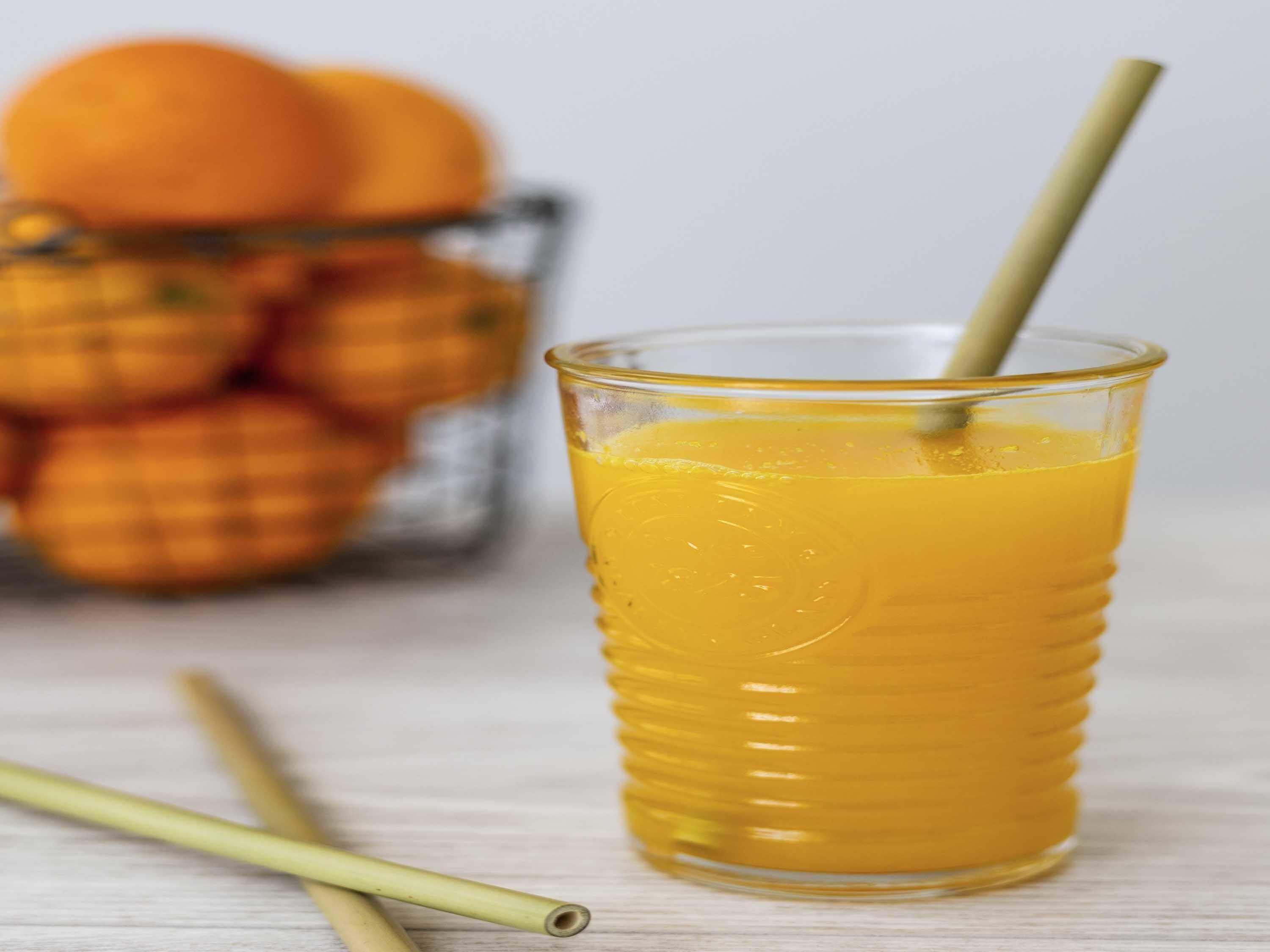 Een glas sinaasappelsap met rietje, een glazen recipiënt met sinaasappels, en twee rietjes op een houten ondergrond.