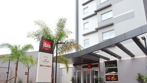 Une entrée d’hôtel moderne, avec une enseigne indiquant la présence d’hôtels ibis, une galerie couverte et un espace paysager agrémenté de palmiers.