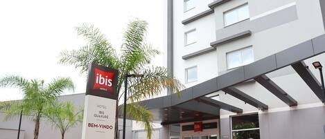 A modern hotel entrance with a sign for ibis hotels, a covered walkway, and a landscaped area with palm trees.