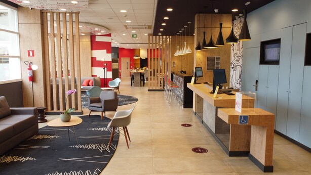 Un espace d’accueil moderne avec un comptoir de réception en bois, un coin salon avec des canapés et des fauteuils, et une grande fenêtre laissant entrer la lumière naturelle.