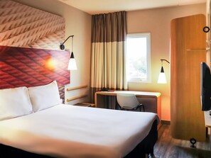 A hotel room with a large bed, a desk with a chair, a TV, and a window with curtains. - Ibis Guaxupe (Guaxupé)