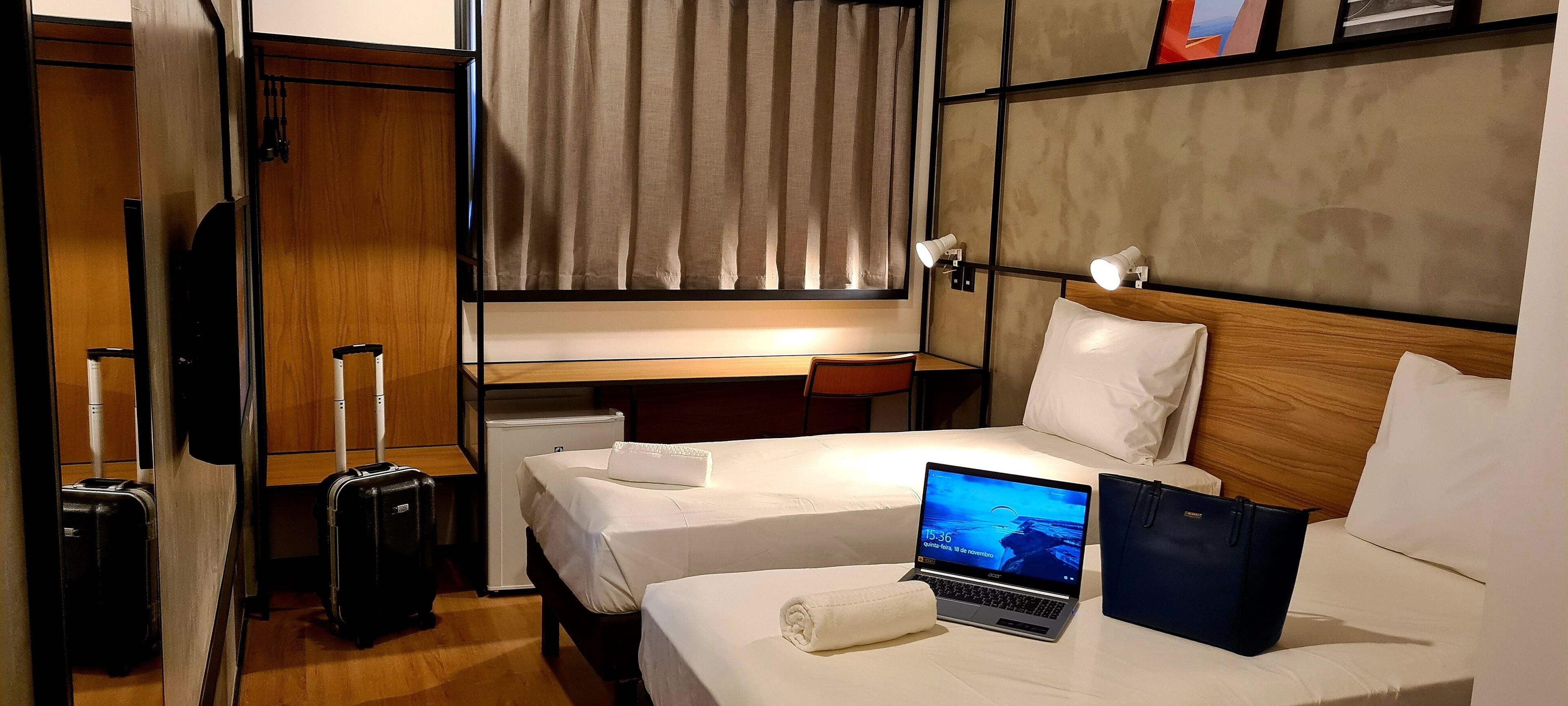 A hotel room with two beds, a desk, a laptop, a suitcase, and a small table.