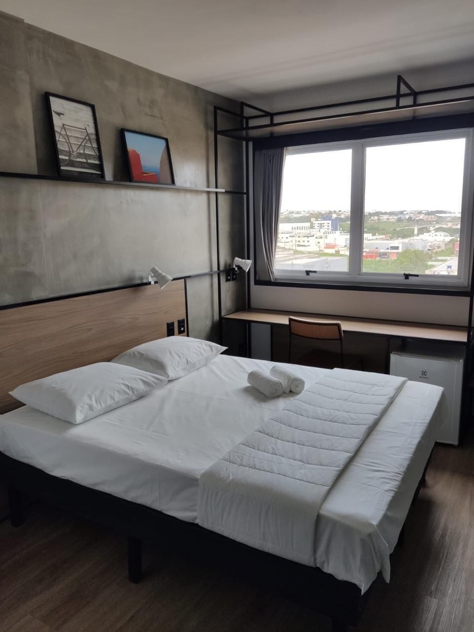 A hotel room with a bed, a desk, a chair, a window with a view, and artwork on the wall.