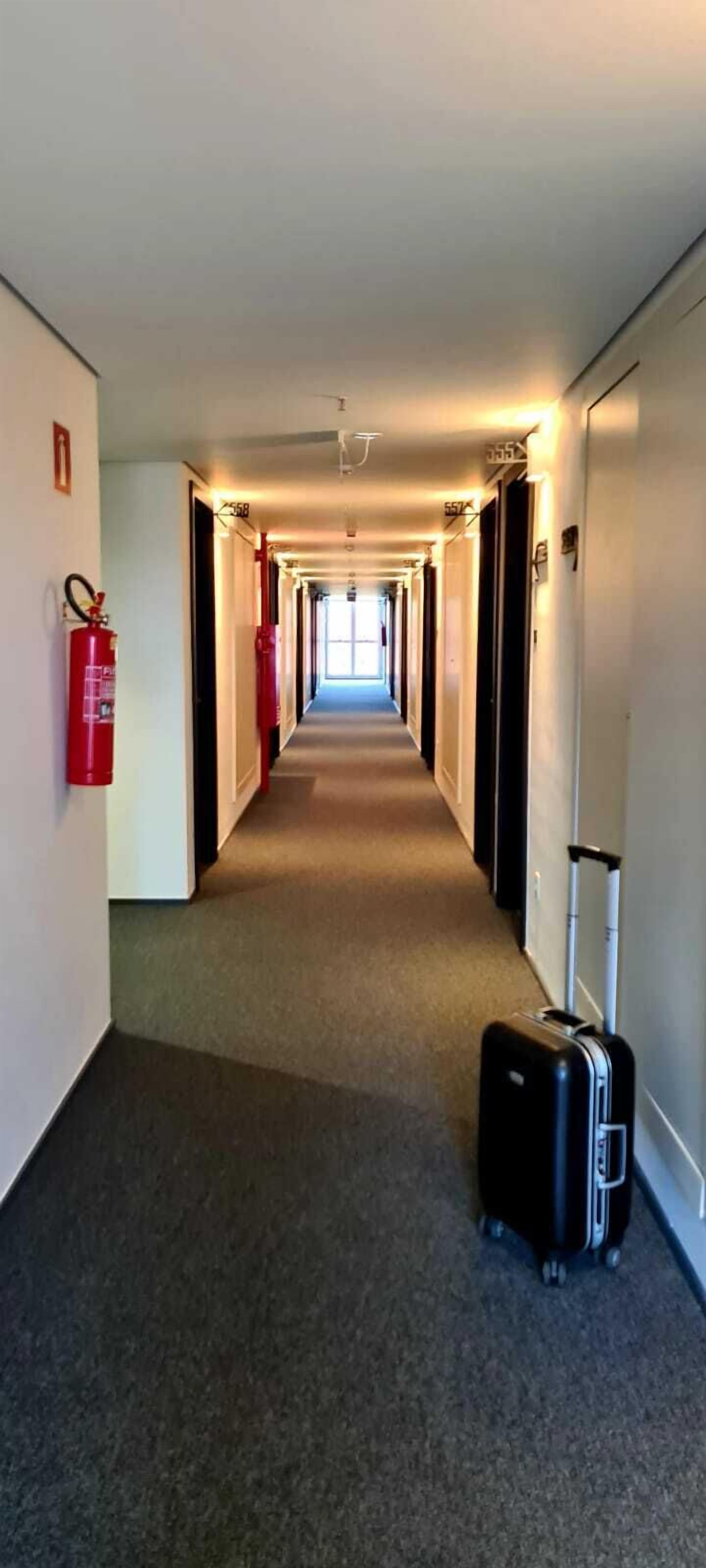 Um corredor de hotel com piso carpete, extintores de incêndio nas paredes e uma mala à direita.