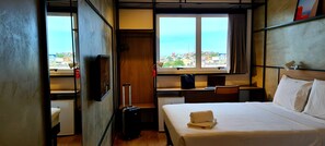 Une chambre d’hôtel avec un lit, un bureau, une chaise, une valise et une vue sur la ville par la fenêtre.
