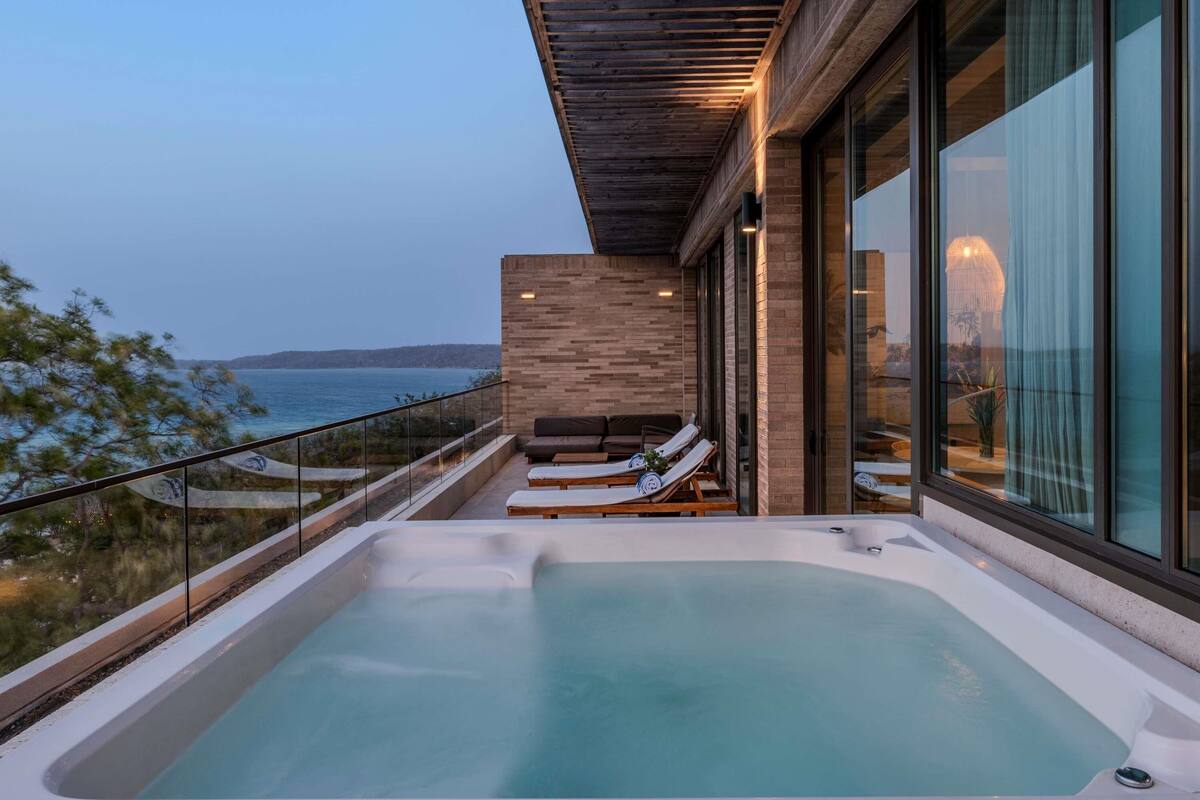 A modern outdoor hot tub area with a view of the sea, a wooden pergola, and a glass balustrade.