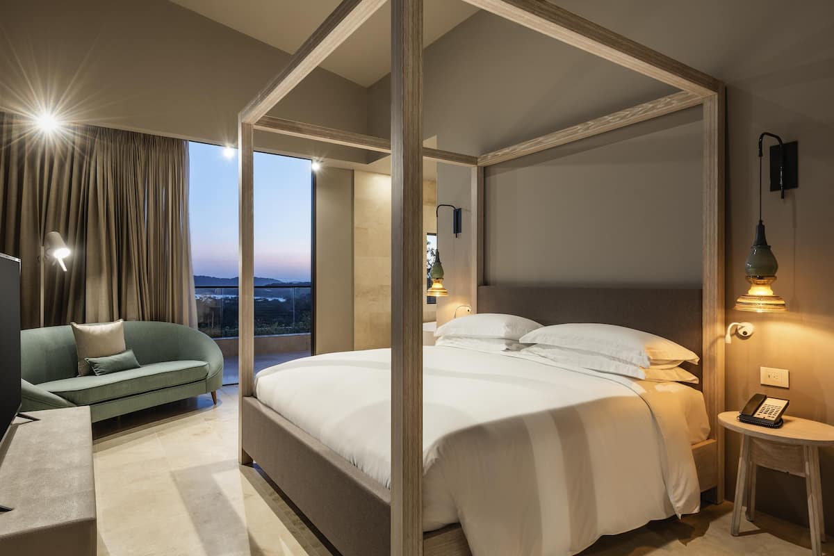 A modern hotel room with a four-poster bed, a sofa, a desk, and a view of the cityscape.