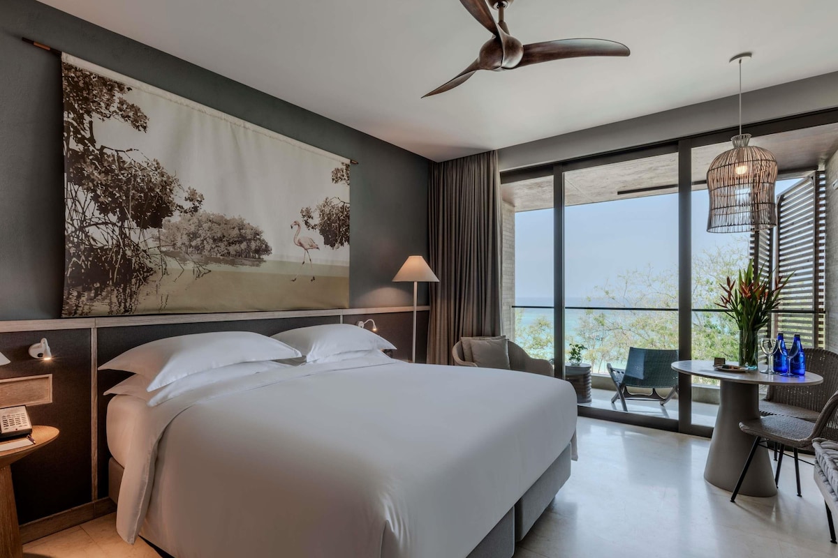 A modern hotel room with a large bed, a desk, a chair, a balcony with a view, and a ceiling fan.
