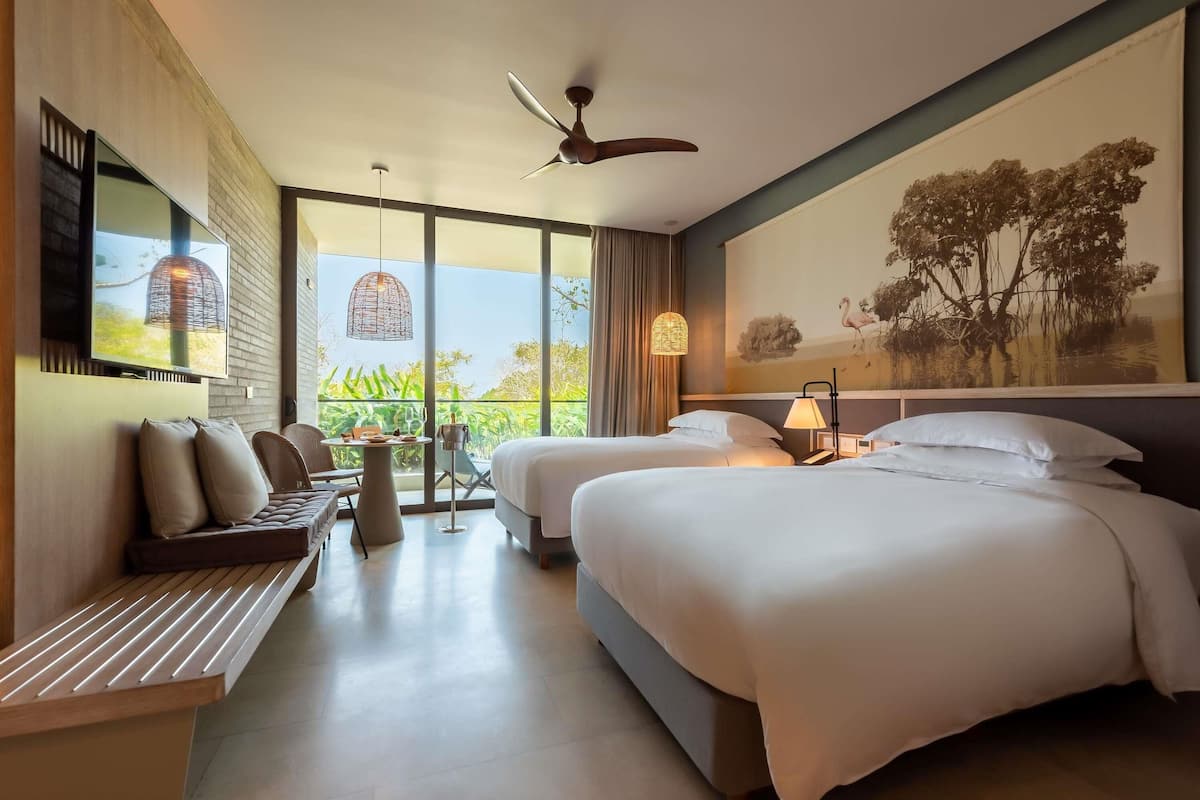 A modern hotel room with two beds, a sitting area with a bench and table, a ceiling fan, and a large window offering a view of greenery.