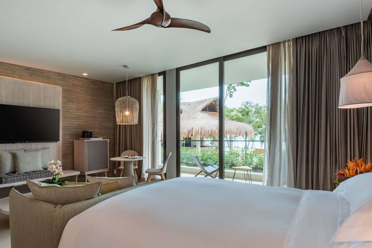 Habitación de hotel moderna con una cama grande, televisión, escritorio y balcón con techo de paja.