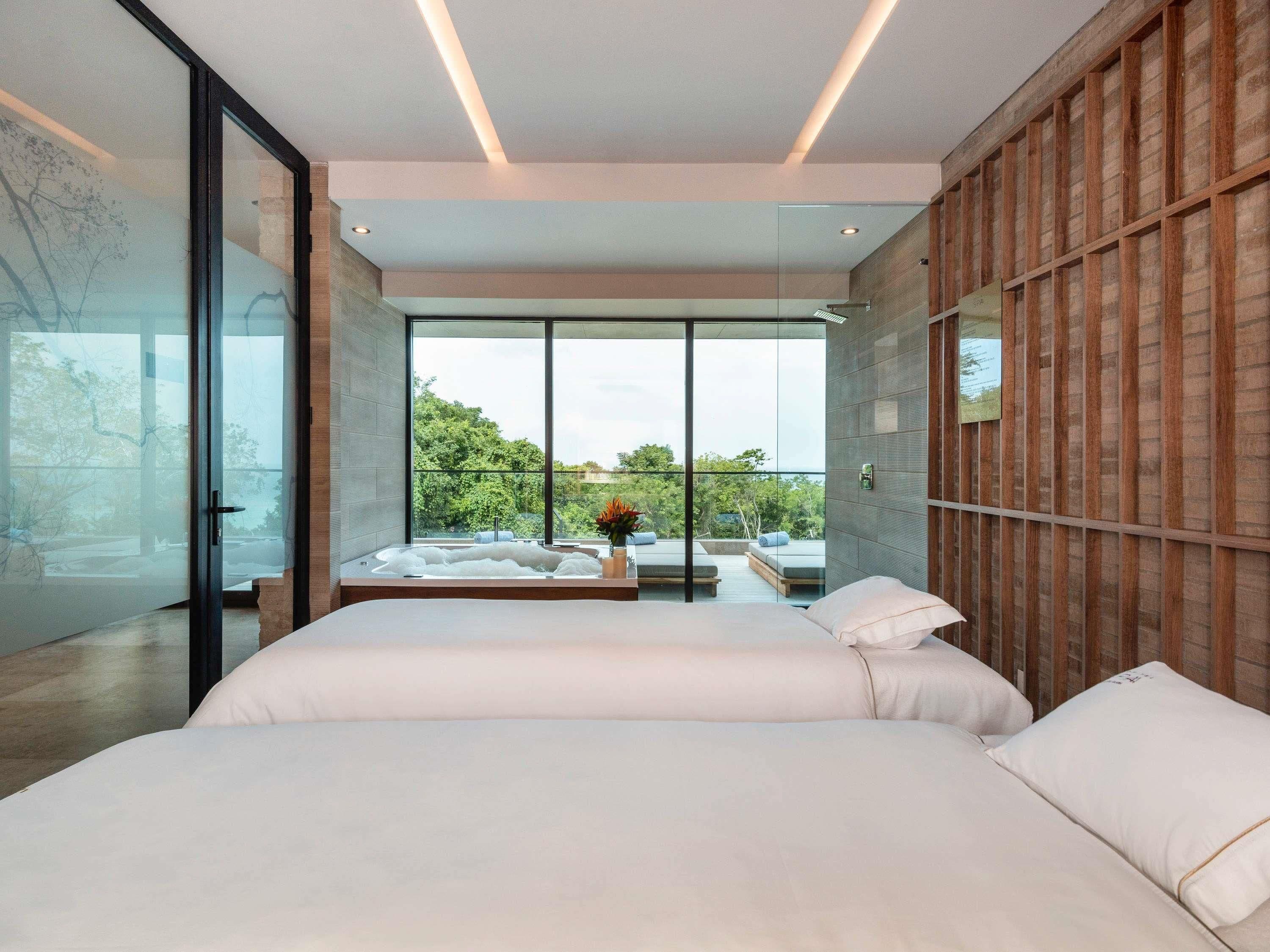 A modern bedroom with a large bed, a glass wall, and a view of greenery outside.