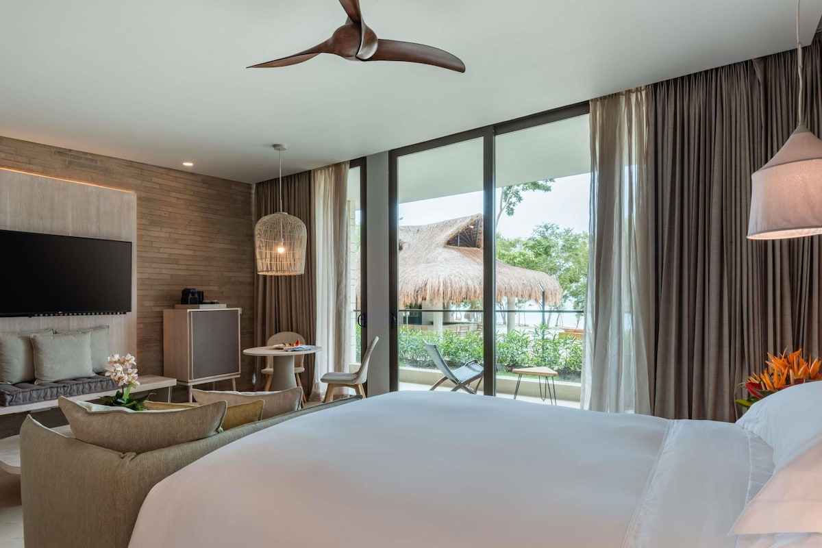 A modern hotel room with a large bed, a TV, a desk, and a balcony with a thatched roof.