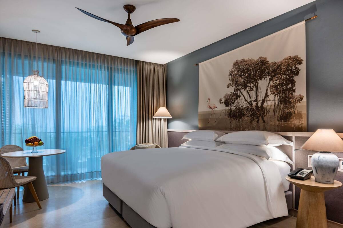 A modern hotel room with a large bed, a desk, a chair, a ceiling fan, and a large window with curtains.
