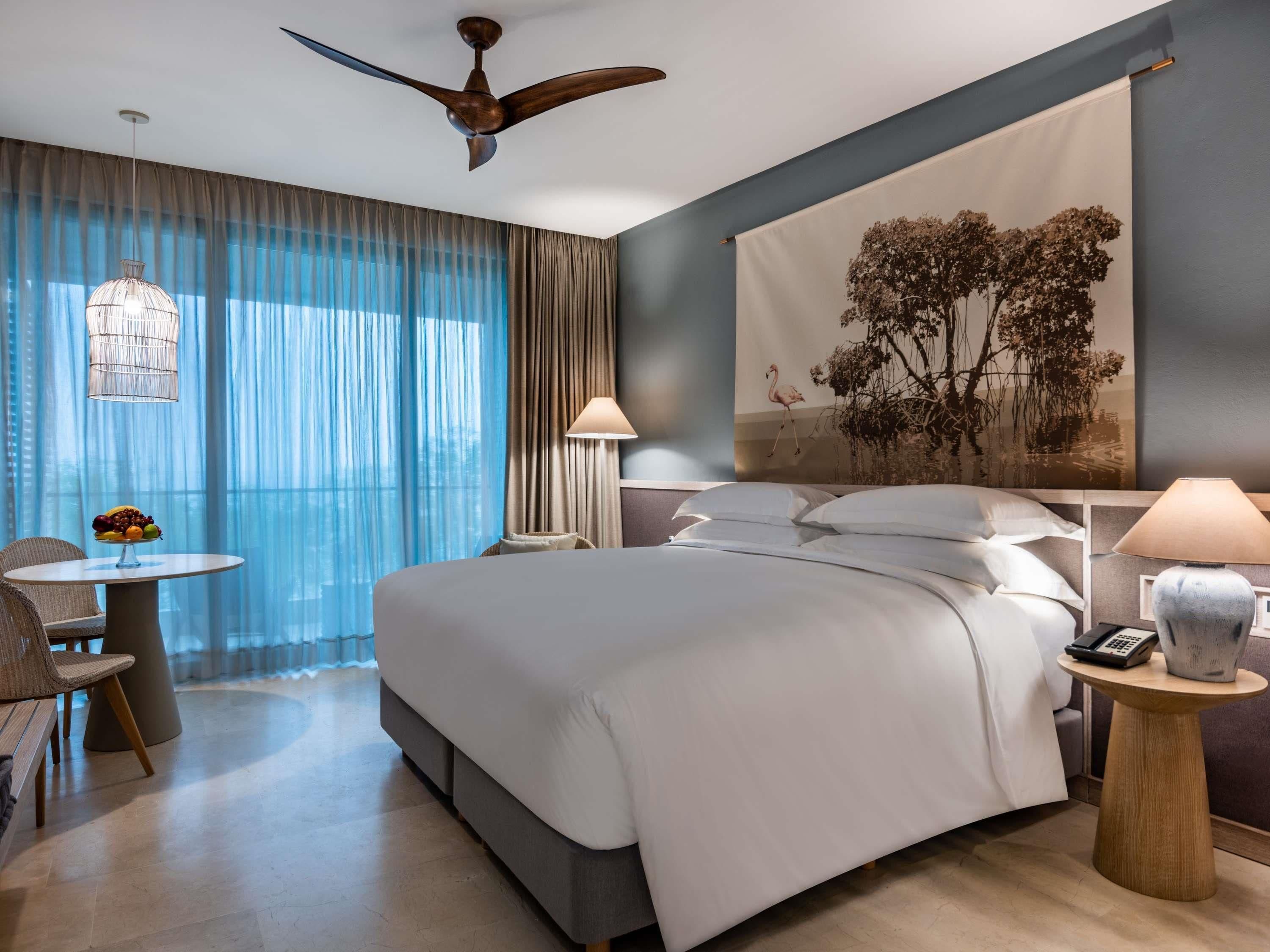A modern hotel room with a large bed, a desk, a chair, a ceiling fan, and a large window with curtains.