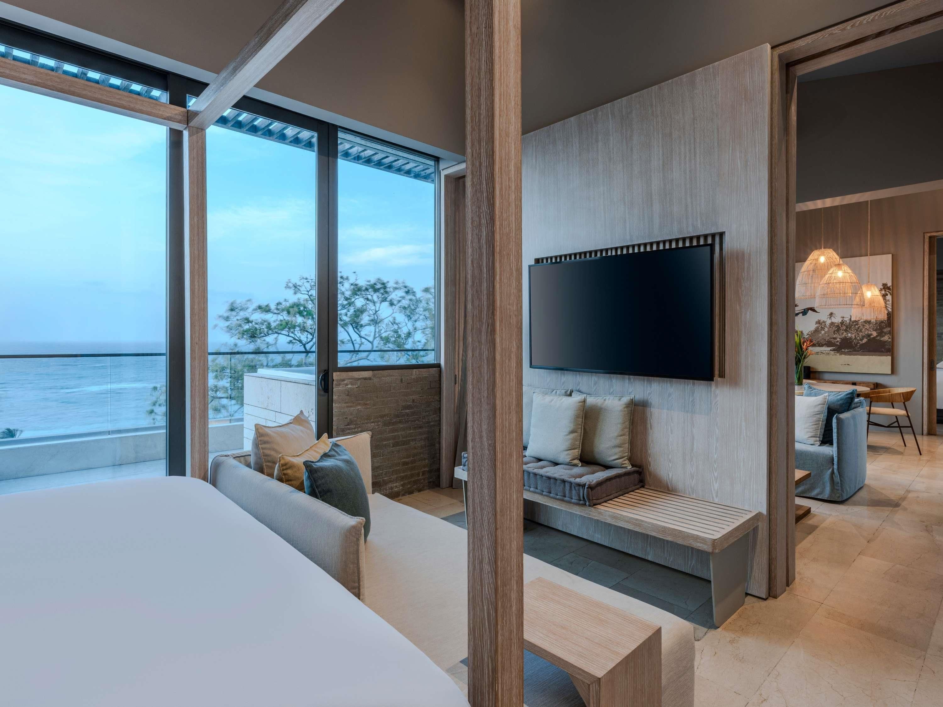A modern bedroom with a large bed, a seating area with cushions, a TV mounted on the wall, and a view of the ocean.