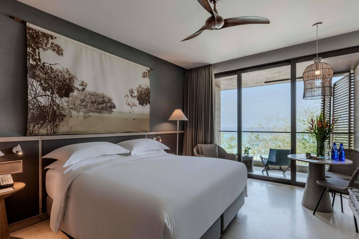 A modern hotel room with a large bed, a desk, a chair, a balcony with a view, and a ceiling fan.