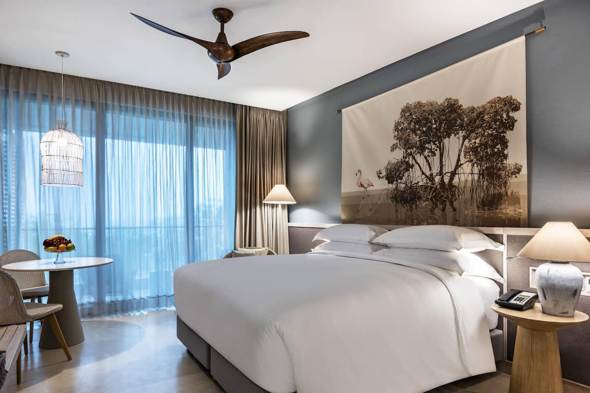 A modern hotel room with a large bed, a desk with a lamp, a chair, and a ceiling fan.