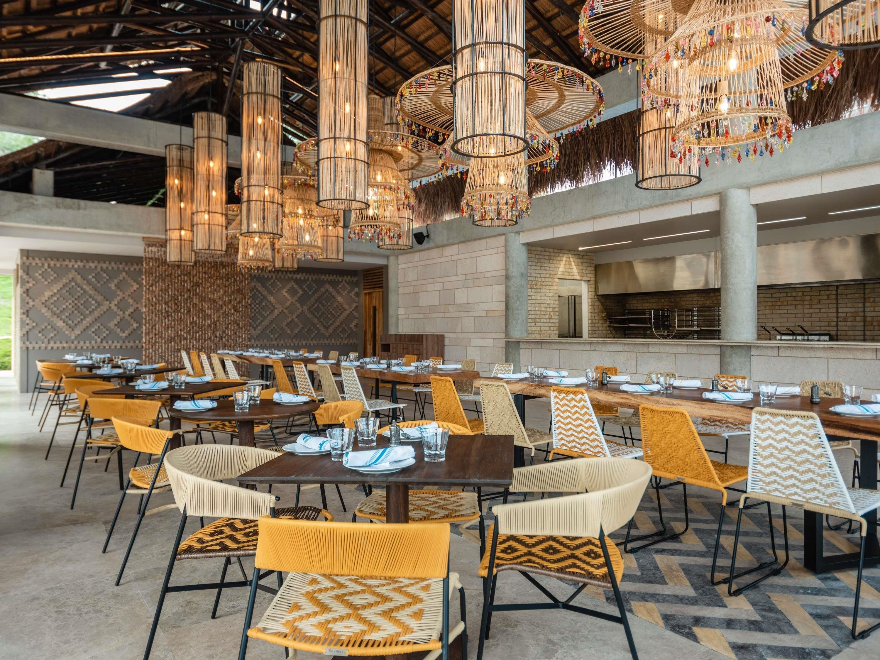 A spacious dining area with multiple tables and chairs, featuring a modern design with exposed brick walls and large hanging light fixtures.