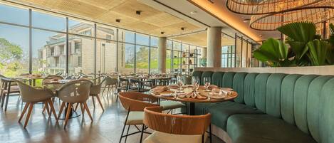 Ein Restaurant mit runden Tischen, grünen Sitzflächen und großen Fenstern mit Blick auf Gebäude und Grünflächen.