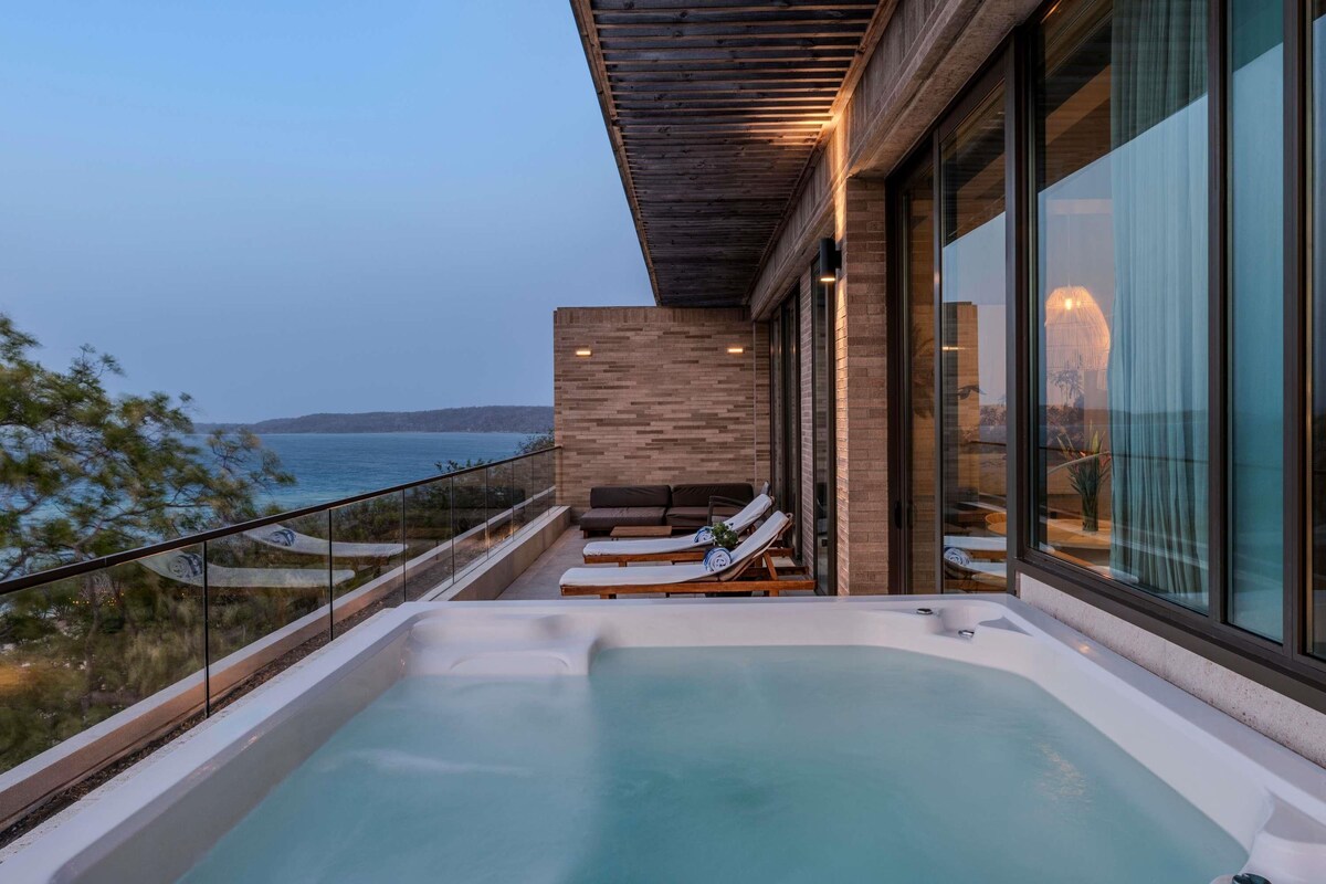 A modern outdoor hot tub area with a view of the sea, a wooden pergola, and a glass balustrade.