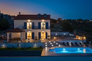 A two-story house with a swimming pool, outdoor seating, and a well-lit balcony.