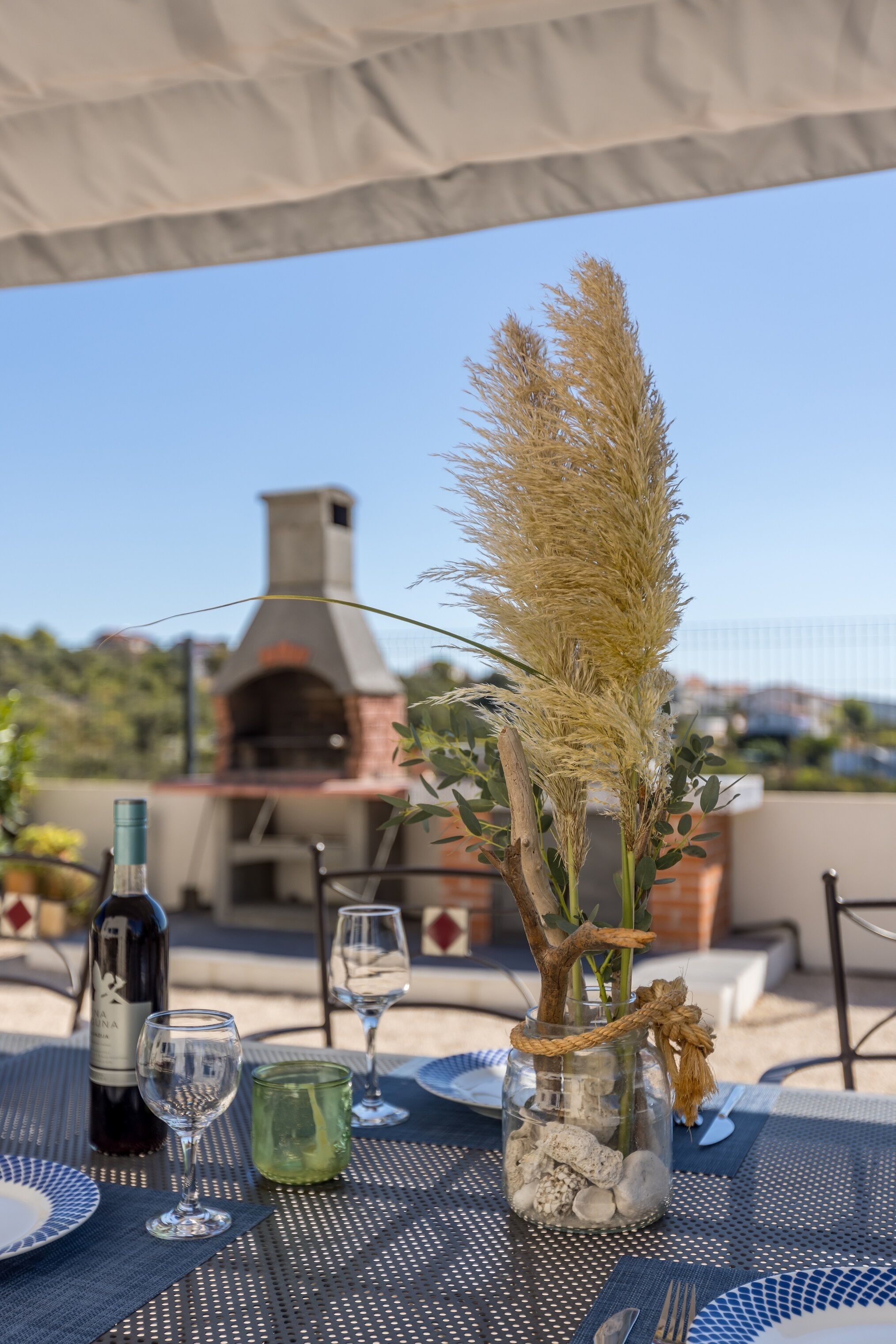 An outdoor dining area with a table set for two, a bottle of wine, and a glass. A decorative vase with dried pampas grass and a branch is on the table. In the background, there is a brick oven and a view of a residential area.