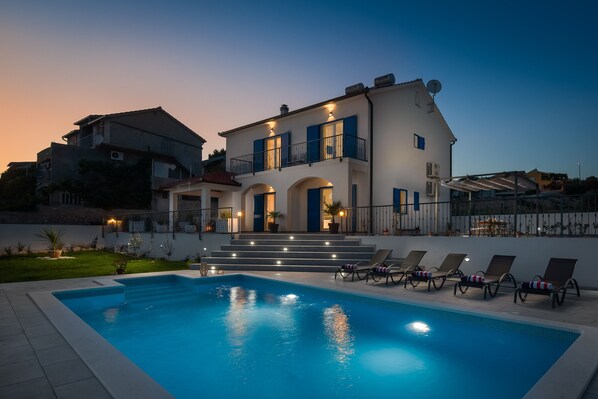 A two-story house with a swimming pool, outdoor seating, and a well-lit entrance.