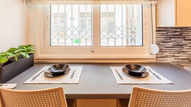 Comedor con una mesa dispuesta para dos personas, una ventana con vistas a edificios y una planta en maceta a la izquierda.