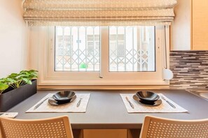 Uma sala de jantar com uma mesa posta para dois, uma janela com vista para edifícios e uma planta em vaso à esquerda.