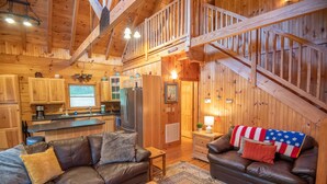 Un acogedor interior de cabaña con detalles de madera, una zona de cocina, una sala de estar con dos sofás de cuero y una escalera.