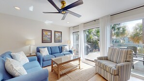 A living room with a blue sofa, a striped armchair, a coffee table, and a ceiling fan. There are two framed pictures on the wall, a window with curtains, and a view of palm trees outside. - Hickory Cove Villa Near Beach, Golf & Pickle Ball at Palmetto Dunes (Hilton Head)