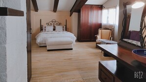 A bedroom with a wooden floor, a bed with a white headboard, a wooden wardrobe, a chair, and a table with a bowl.